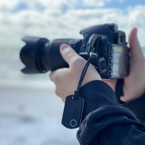 Hand holding a camera with a dog tag attached, blurred water and sky in the background