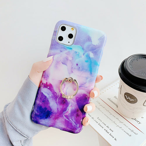 Hand holding a phone with a purple and blue marbled case and ring holder, with a coffee cup and book in the background.